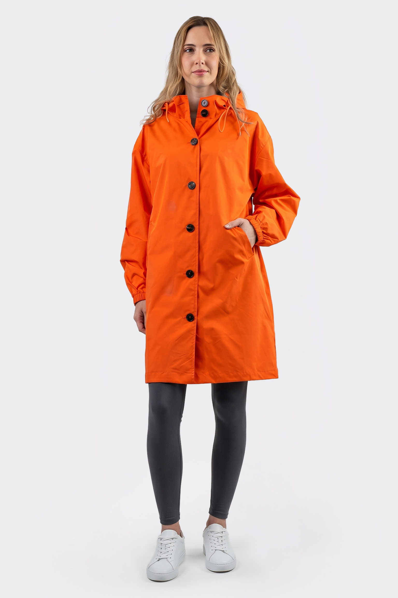 A person stands facing forward wearing the Icon Windbreaker Rain Jacket in orange, paired with black leggings and white sneakers, against a plain white background.