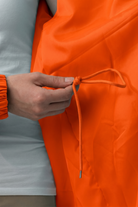 A person wearing a white shirt holds the drawstring of the Icon Windbreaker Rain Jacket in orange, with an orange scrunchie on their wrist.