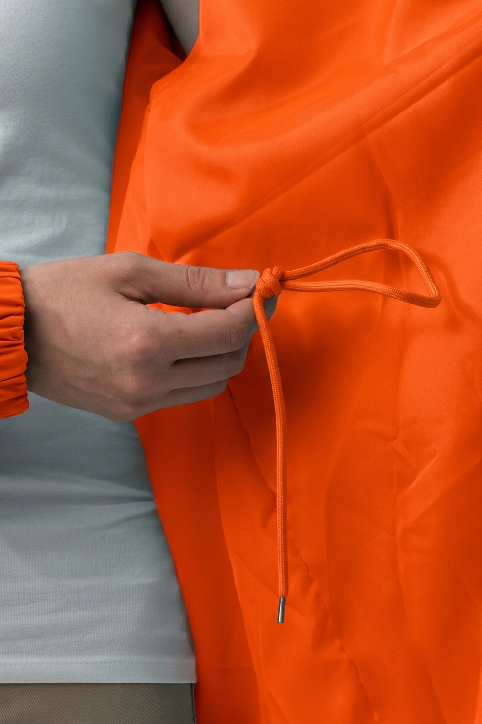A person wearing a white shirt holds an orange drawstring tied in a bow on The Water Resistant Hooded Oversized Windbreaker Rain Jacket. Only the hand, part of the arm, and torso are visible.