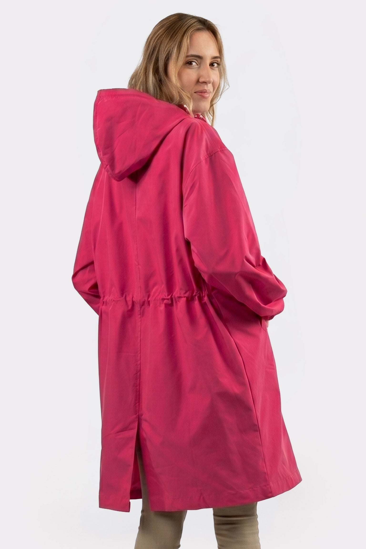 A person with light brown hair is wearing The Water Resistant Hooded Oversized Windbreaker Rain Jacket in bright pink. With an adjustable waist and hands in pockets, they stand against a plain light background, back slightly turned.