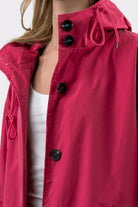 A person wearing the Icon Windbreaker Rain Jacket in bright pink with black buttons and a hood over a white top. The image displays the upper torso; part of the face is out of frame.