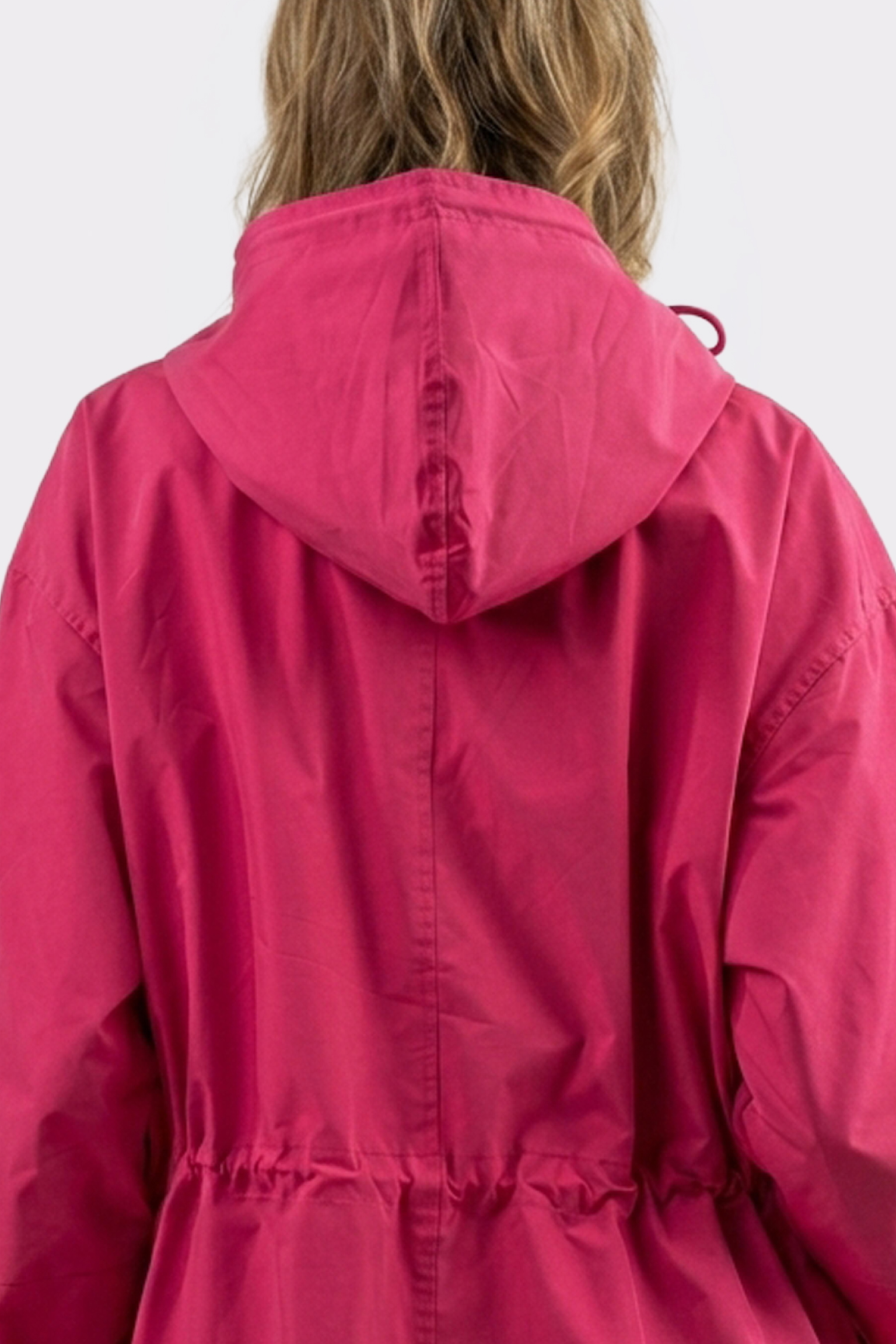 A person is seen from behind wearing The Water Resistant Hooded Oversized Windbreaker Rain Jacket in bright pink, with the hood up partially covering their light brown hair and a drawstring cinching the waist.
