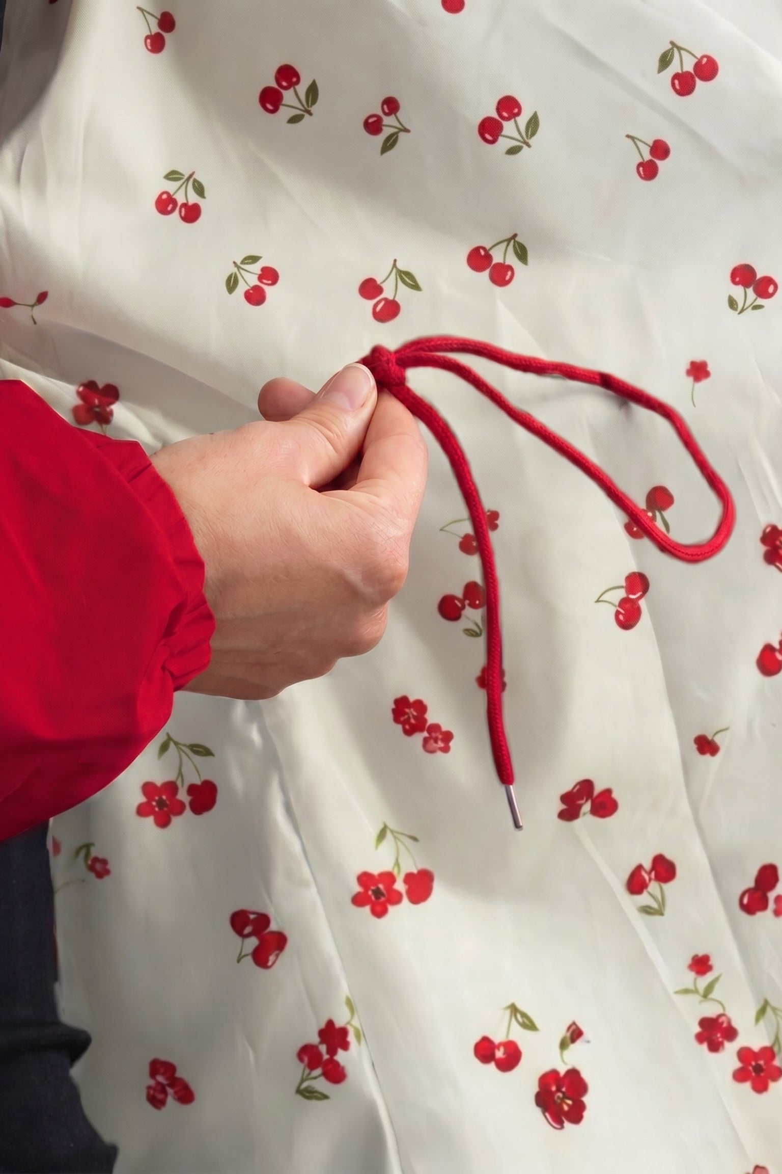 A person wearing a red sleeve holds the red drawstring of the Icon Windbreaker Rain Jacket against white fabric featuring a cherry and flower print.