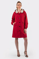 A model wears the Icon Windbreaker Rain Jacket, a red knee-length coat with buttons, pockets, and a hood, styled with black open-toed heels against a plain white background.