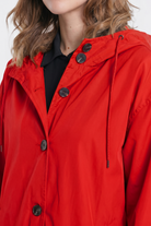 A person with wavy light brown hair wears a bright red Icon Windbreaker Rain Jacket with black buttons, drawstrings, a hood, and layers it over a black top.