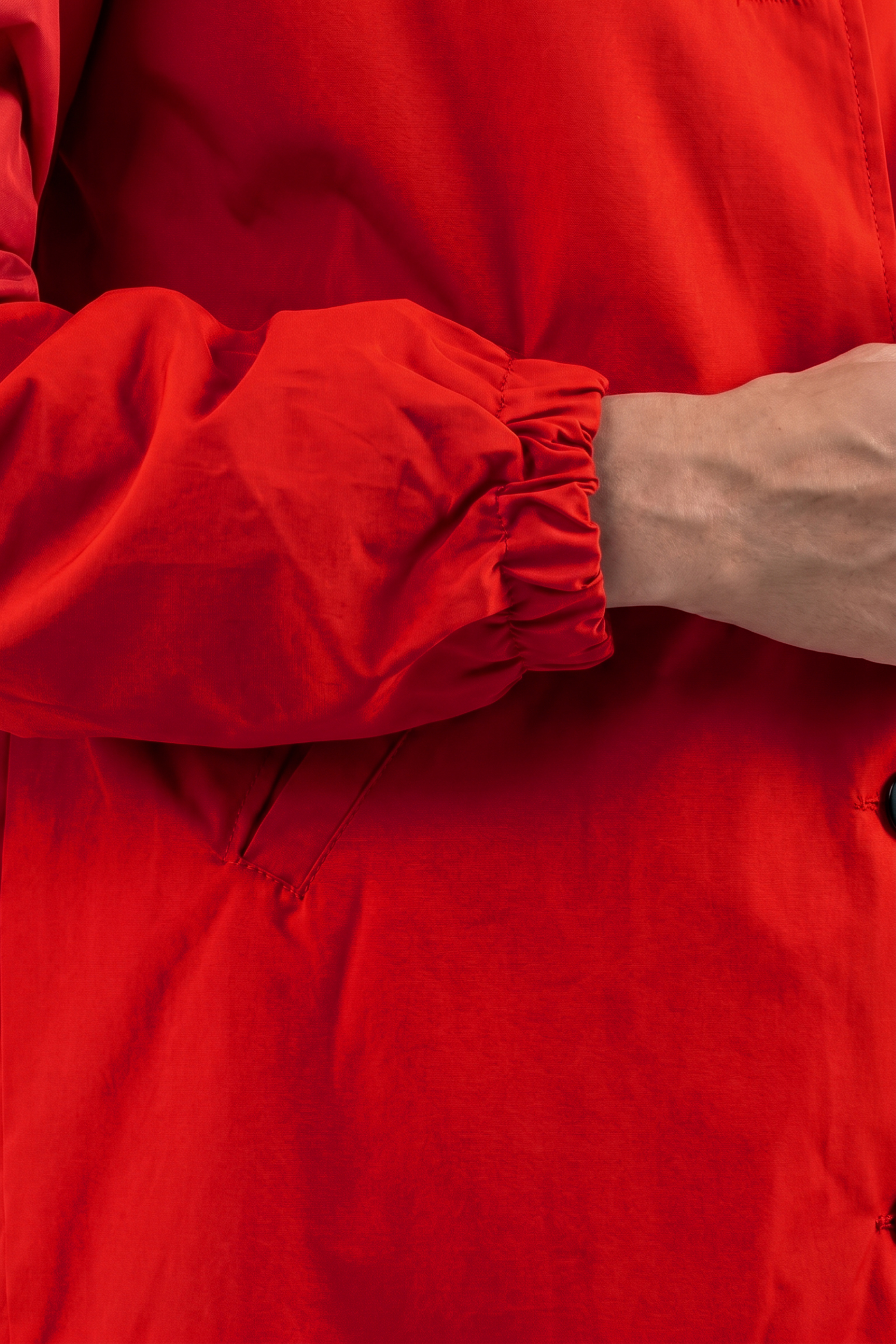Wearing a bright red Icon Windbreaker Rain Jacket with elastic cuffs, a person crosses their arm over their body. Only the torso and part of one arm are visible in the image.