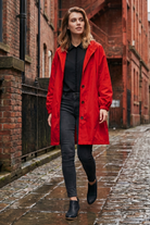 A woman wearing the Icon Windbreaker Rain Jacket, a black shirt, dark jeans, and black shoes walks on a wet cobblestone street lined with brick buildings.