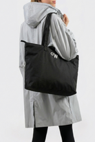 A person in a light gray hooded coat and black leggings holds the large black Urban Slate Waterproof Bag with "CW" printed on it, standing against a plain, light-colored background.
