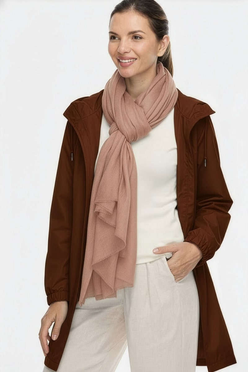 A woman stands smiling with her left hand in her pocket, her hair tied back, wearing a brown jacket, off-white top, beige pants, and the looselyboho Sandstone Dune 100% Cashmere Scarf—a chic touch for winter layering. The background is plain and light.