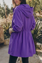 A woman stands outdoors wearing a long, purple Icon Rain Windbreaker Jacket with a gathered waist and hood. She is facing away, looking to the side, with greenery and rocks in the background.