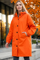 A woman in the Icon Windbreaker Rain Jacket stands outside by modern glass buildings and autumn trees with orange leaves.