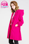 A person with shoulder-length wavy hair is wearing a bright pink, hooded coat made of lightweight water-resistant fabric with black buttons. They have their hands in the coat's pockets and are looking down. The BARBARA PINK WINDBREAKER by looselyboho is labeled in the top left corner.