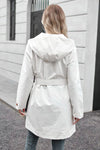 A person with long hair, seen from behind, wearing a BELTED FROST WHITE WINDBREAKER by looselyboho and blue jeans stands in front of a modern building with light-colored walls and a dark doorway.