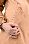 Close-up of a person's hand pulling on a pocket flap of a tan-colored, stylish rain-jacket. The person is wearing the PEACH FUZZ WINDBREAKER by looselyboho, made from lightweight water-resistant material, and the sleeve has an elastic cuff. The background is plain white.