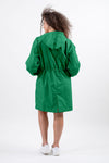 A person with curly hair faces away from the camera, wearing an emerald green EMERALD GREEN WINDBREAKER by The Windbreaker Jacket made of water-resistant material and white sneakers. The coat has an elastic waistband, an adjustable hood, and the background is plain white.