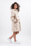 A person with curly hair stands against a plain white background, wearing a BISCUIT BEIGE WINDBREAKER by looselyboho with an adjustable hood and hands in pockets. They are looking to the side and are dressed casually with white sneakers.