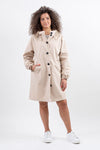 A woman with curly hair is standing against a plain white background. She is wearing the looselyboho BISCUIT BEIGE WINDBREAKER, beige and knee-length with black buttons, slightly open at the collar. The lightweight, water-resistant coat features an adjustable hood. She has one hand in her pocket and is wearing white sneakers.