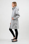 A woman is standing sideways, dressed in a light gray hooded **ICE GRAY WINDBREAKER** made by **looselyboho**, crafted from water-resistant material with an adjustable hood. She wears black leggings and black flats. Her long sandy blonde hair flows freely in a neutral studio setting with a plain white background.